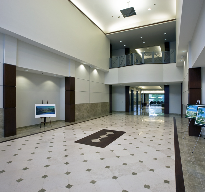 Spacious modern lobby with high ceilings, tiled floor, a painting on an easel, and framed pictures. Large windows in the background provide natural light. Balcony overlooks the lobby area.