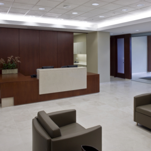 Modern reception area with a wooden desk and two beige chairs. Room features a light marble floor and recessed lighting. A conference room with a glass wall is visible in the background.