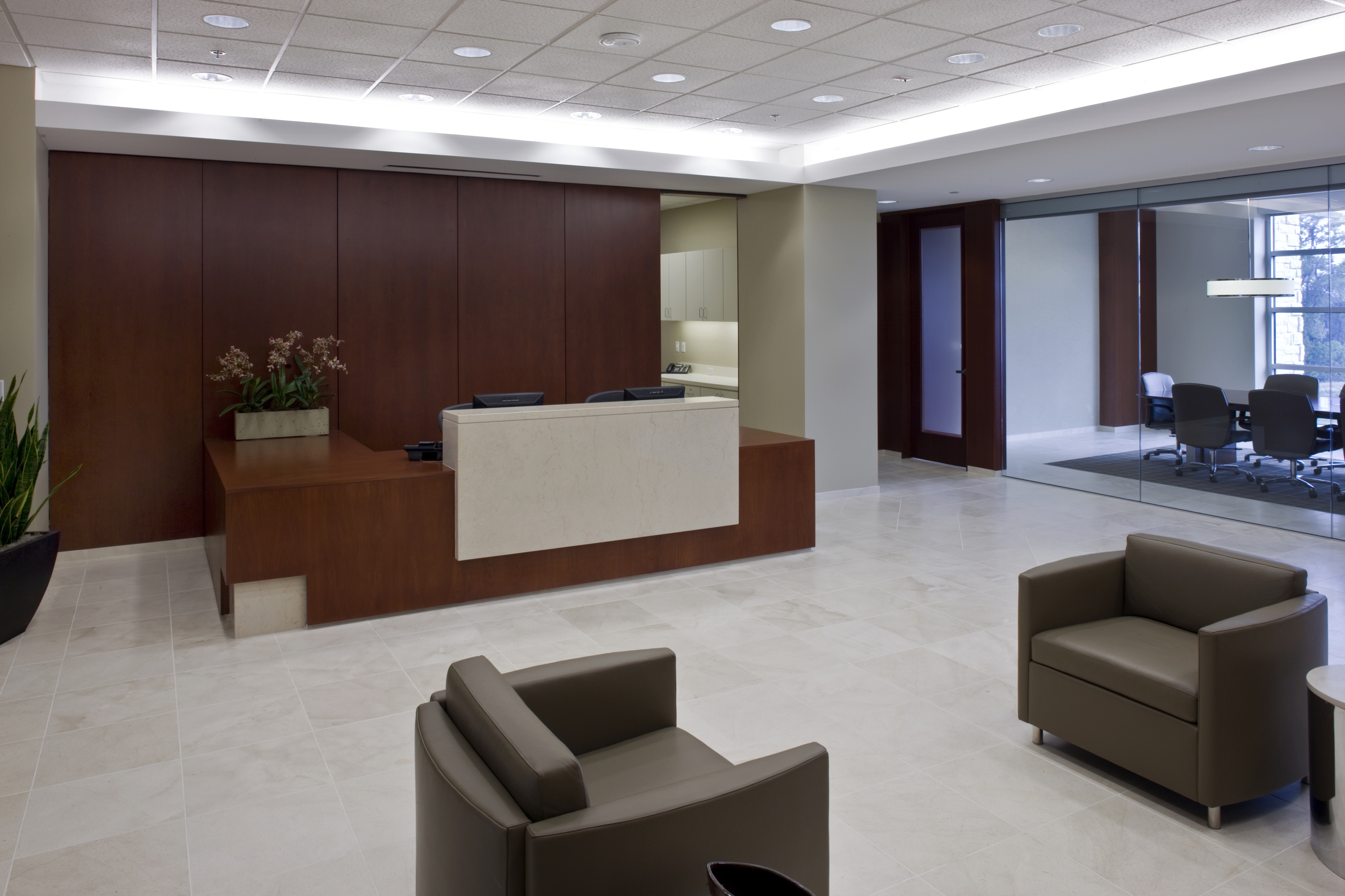 Modern reception area with a wooden desk and two beige chairs. Room features a light marble floor and recessed lighting. A conference room with a glass wall is visible in the background.