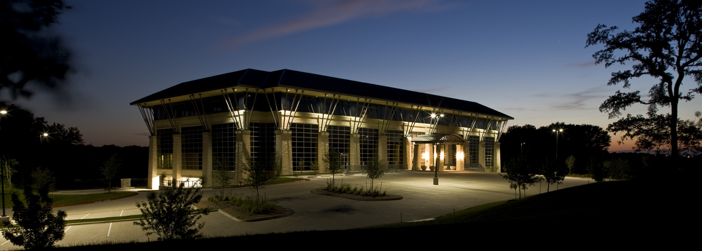 A large modern building with extensive glass windows is illuminated at dusk, surrounded by trees and a mostly empty parking lot.