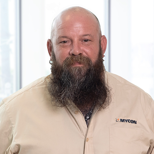A man with a full beard, wearing a beige work shirt labeled "mycon," smiling and standing in an office setting with windows in the background.