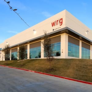 A large, modern warehouse with the letters "wrlg" in red on the upper corner of the building. The structure is located on a grassy incline against a clear blue sky.