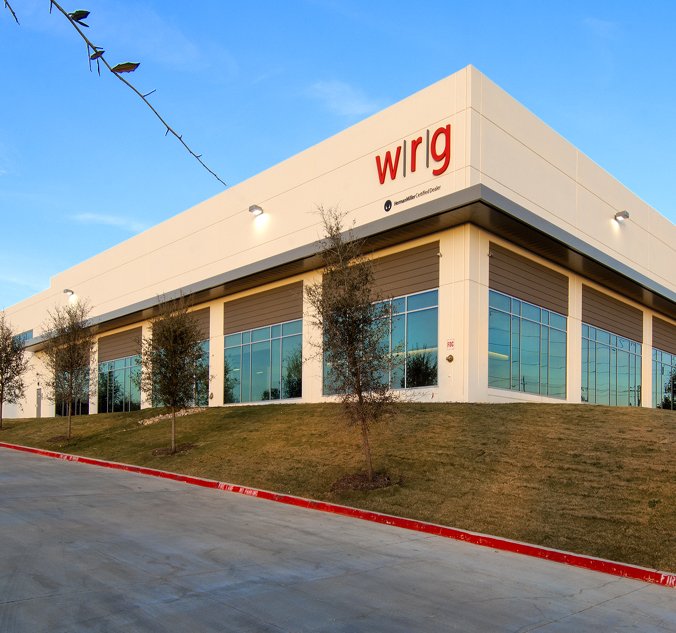 A large, modern warehouse with the letters "wrlg" in red on the upper corner of the building. The structure is located on a grassy incline against a clear blue sky.