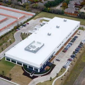 Aerial view of a large, white-roofed industrial building surrounded by a parking lot with multiple cars, located near a residential area and storage units.