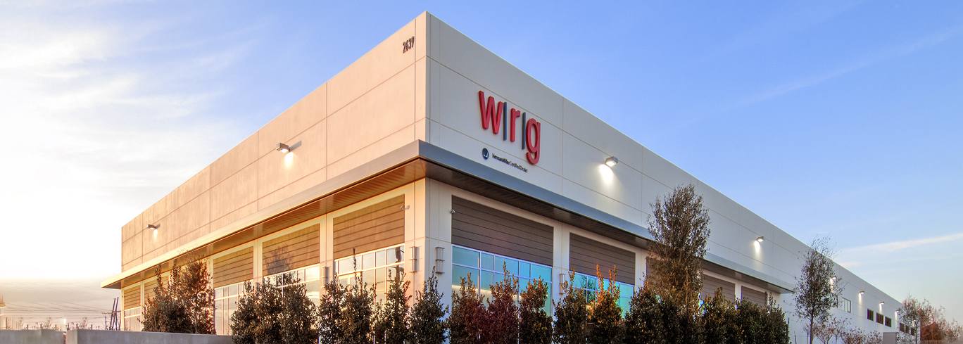 A large modern warehouse building with "WITG" written on the side, surrounded by trees, under a clear blue sky.