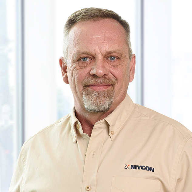 A middle-aged man with a goatee, wearing a beige shirt with a mycon logo, stands smiling in a bright, window-lined office.