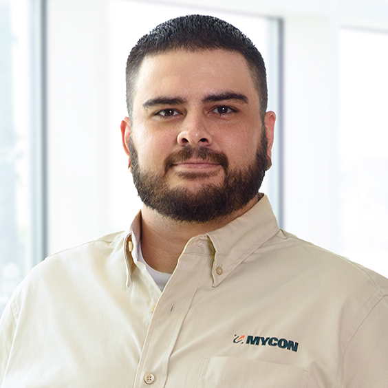 A man with a beard in a beige shirt with "mycon" logo, standing confidently in front of a windowed office background.