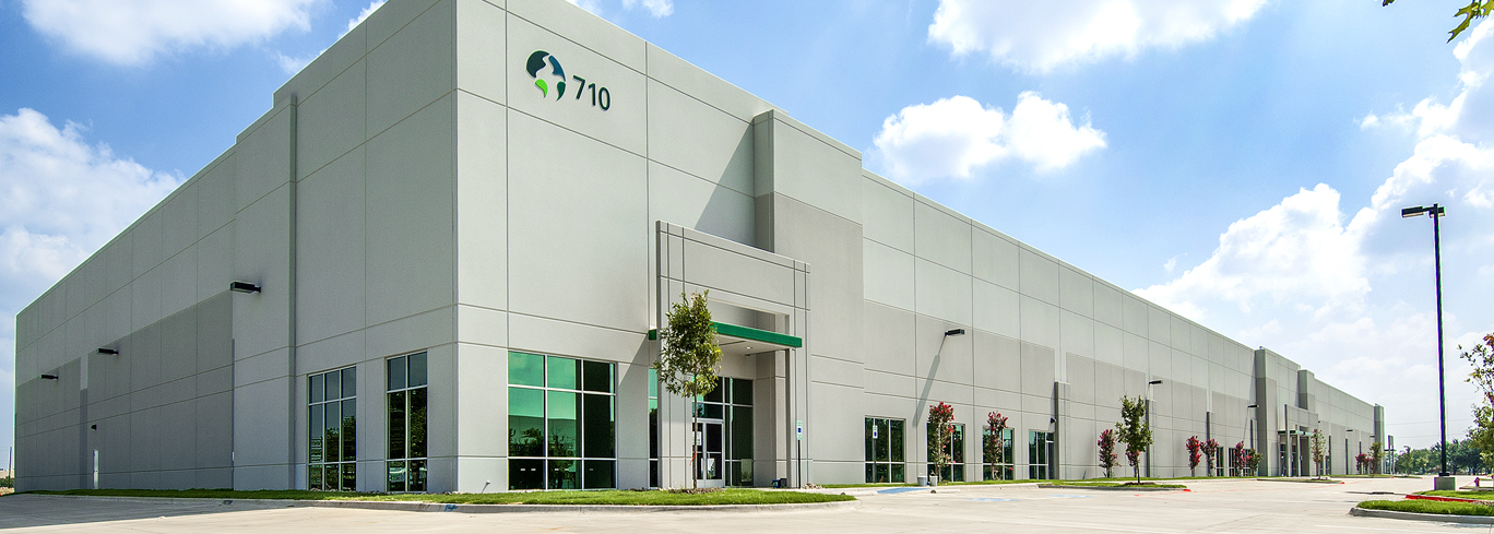 A large, modern industrial building with light gray exterior and green accents, labeled "710," set under a clear sky with a few cars and trees around the perimeter.