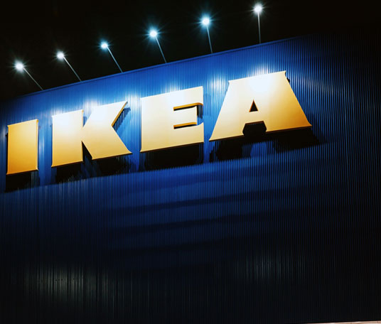A large yellow IKEA sign is displayed on a blue, ribbed wall with bright lights illuminating it from above.