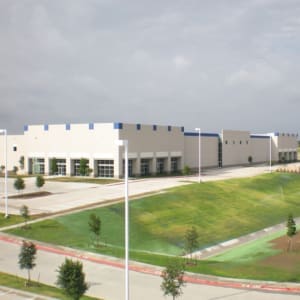 A large, white industrial building with blue accents is shown. The building is surrounded by well-maintained grassy areas and a paved parking lot. The sky is overcast.