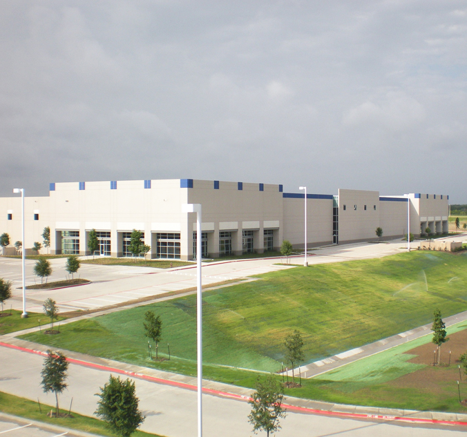 A large, white industrial building with blue accents is shown. The building is surrounded by well-maintained grassy areas and a paved parking lot. The sky is overcast.