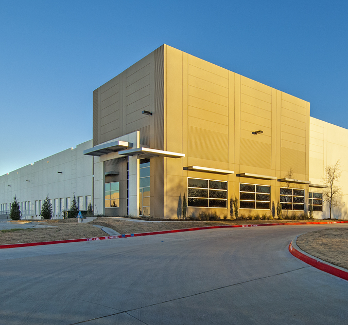 A large, modern industrial warehouse with beige walls and large windows. The building is surrounded by a concrete driveway and sparse landscaping against a clear blue sky.
