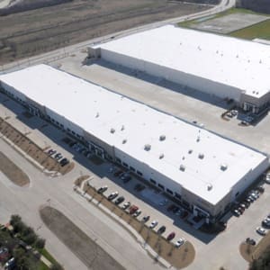 Aerial view of two large industrial buildings with white roofs and multiple parked cars surrounding them in a business area.