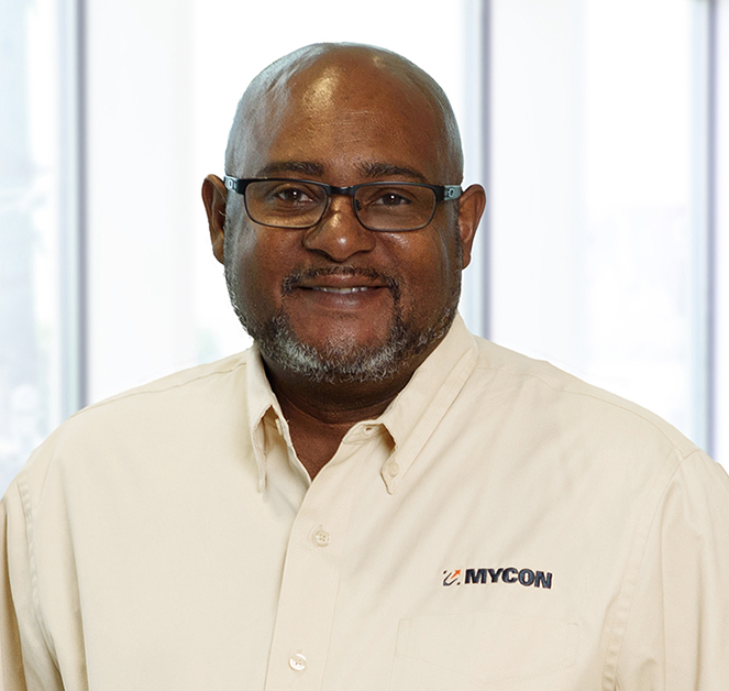 A man wearing glasses and a cream-colored button-up shirt with "MYCON" logo stands smiling in front of a windowed background.