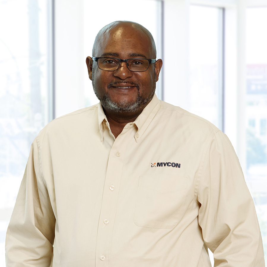 A man wearing glasses and a cream-colored button-up shirt with "MYCON" logo stands smiling in front of a windowed background.