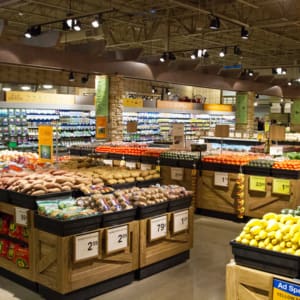 A grocery store produce section with various fruits and vegetables displayed in wooden bins, including tomatoes, potatoes, and squash. Signs with prices are visible on the bins.