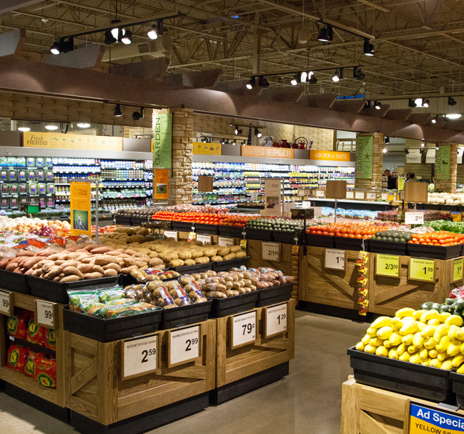 A grocery store produce section with various fruits and vegetables displayed in wooden bins, including tomatoes, potatoes, and squash. Signs with prices are visible on the bins.