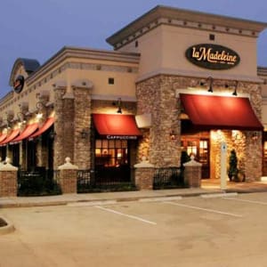 Exterior view of a la Madeleine French Bakery & Café at dusk, featuring stone facade, red awnings, and a lit sign with the café's name.