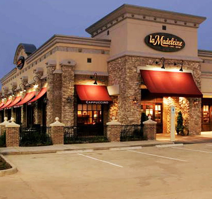 Exterior view of a la Madeleine French Bakery & Café at dusk, featuring stone facade, red awnings, and a lit sign with the café's name.