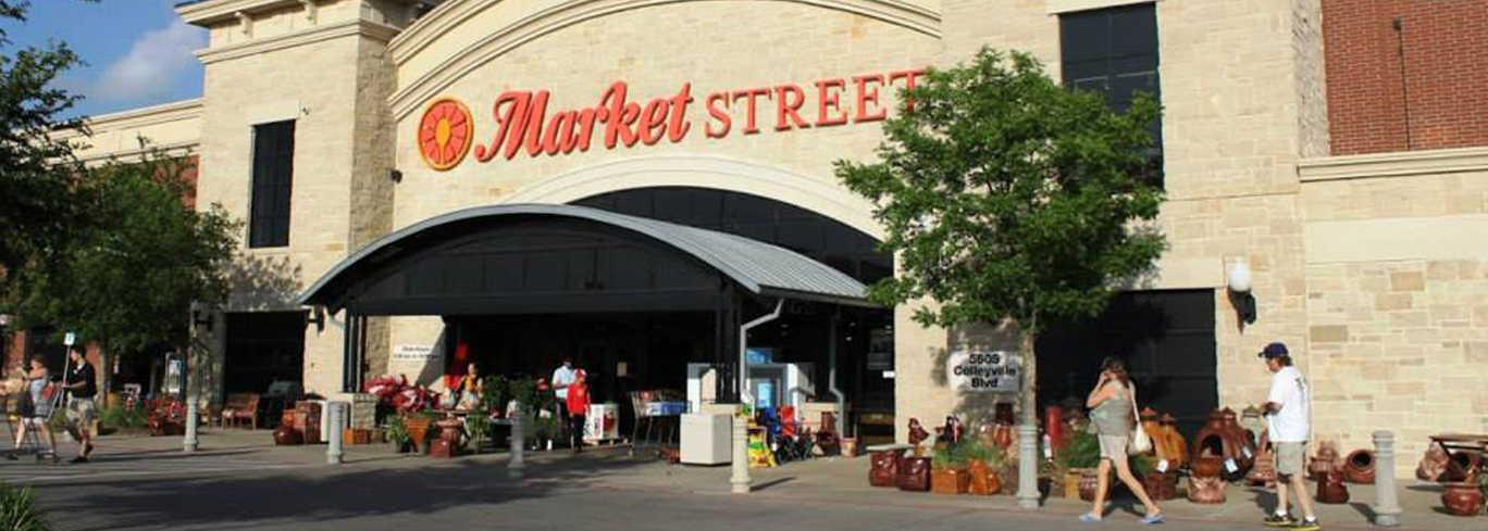 The front entrance of a market, marked "Market Street," features various potted plants and shoppers walking in and out. The building has a mix of brick and stone exterior with a metal awning.