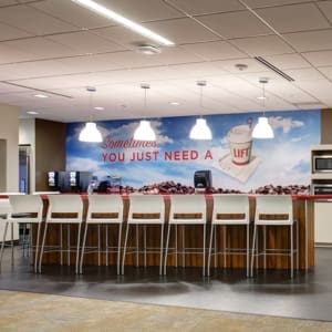 Long counter with stools in a break room, featuring a mural of a coffee cup and text "Sometimes YOU JUST NEED A LIFT." Overhead lights illuminate the counter area, which has coffee machines and microwaves.