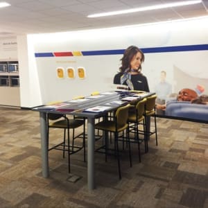 A modern office space with high tables and stools. A large wall graphic shows a smiling flight attendant interacting with a passenger. The floor is carpeted with a neutral pattern.