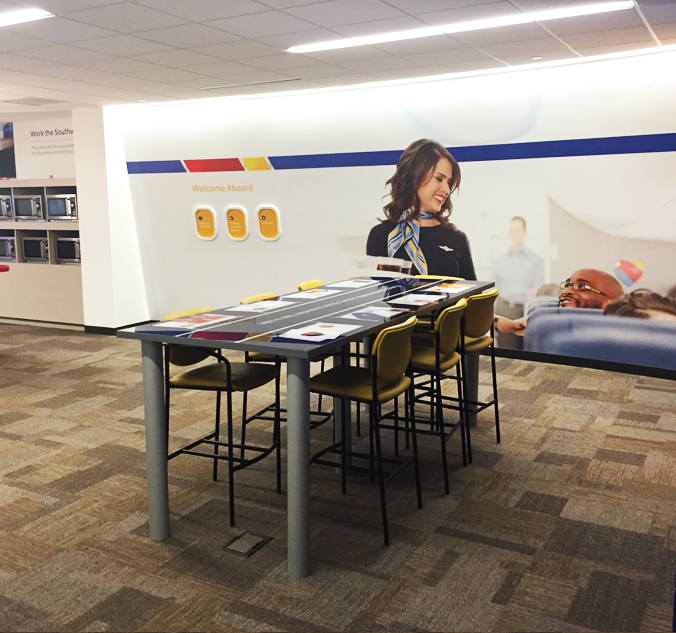 A modern office space with high tables and stools. A large wall graphic shows a smiling flight attendant interacting with a passenger. The floor is carpeted with a neutral pattern.