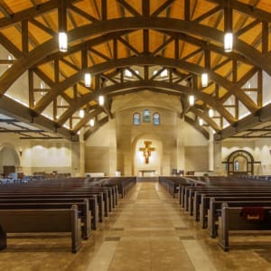 Interior of a modern church with wooden beams, pendant lighting, and rows of empty pews leading to an altar with a large crucifix. Stained glass windows are visible above the altar.