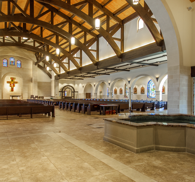 Interior of a modern church with a high wooden beam ceiling, pews, stained glass windows, religious statues, and a baptismal font in the foreground.