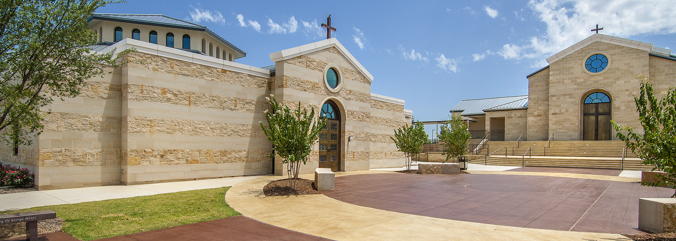 A modern building complex with beige stone walls and multiple crosses on top. The paths are clean, and there are trees planted near the buildings. The sky is clear with a few scattered clouds.