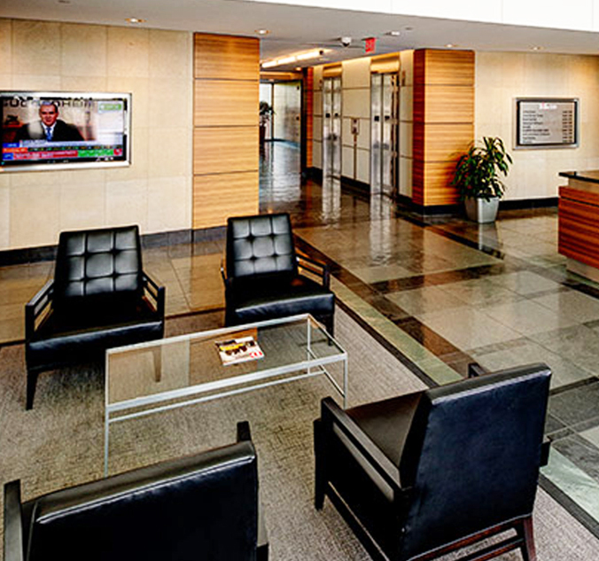 A modern office lobby with four black leather chairs around a glass table, a TV on the wall, a potted plant, and an elevator area in the background.