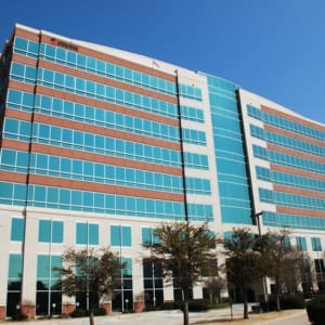 A multi-story office building with a mix of brick and blue-tinted glass windows stands under a clear blue sky. Trees are visible in the foreground.
