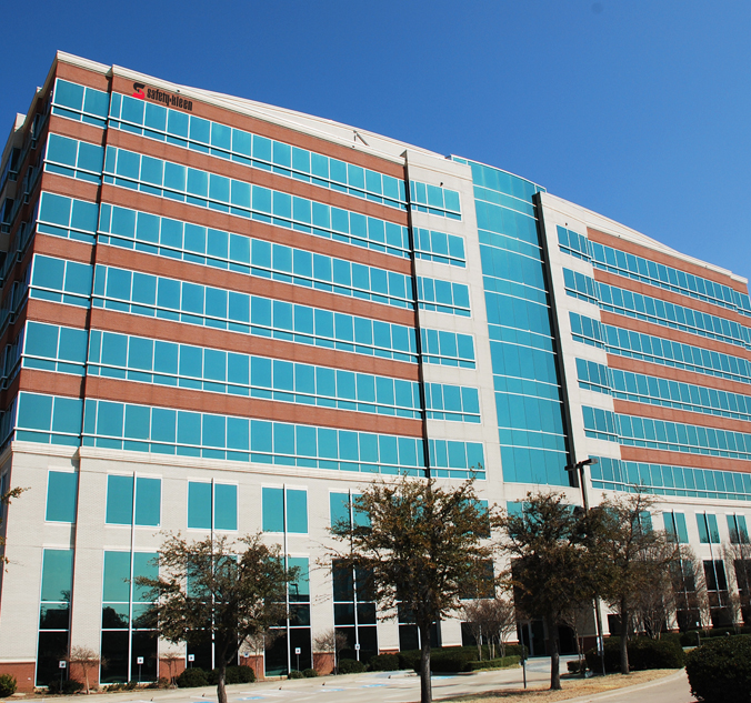 A multi-story office building with a mix of brick and blue-tinted glass windows stands under a clear blue sky. Trees are visible in the foreground.