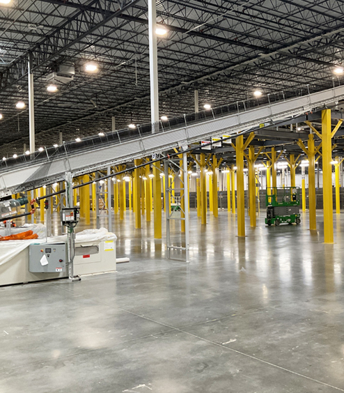 A large warehouse interior featuring a conveyor system, yellow support columns, and various machinery. The space is brightly lit, clean, and organized, indicating a functional industrial facility.