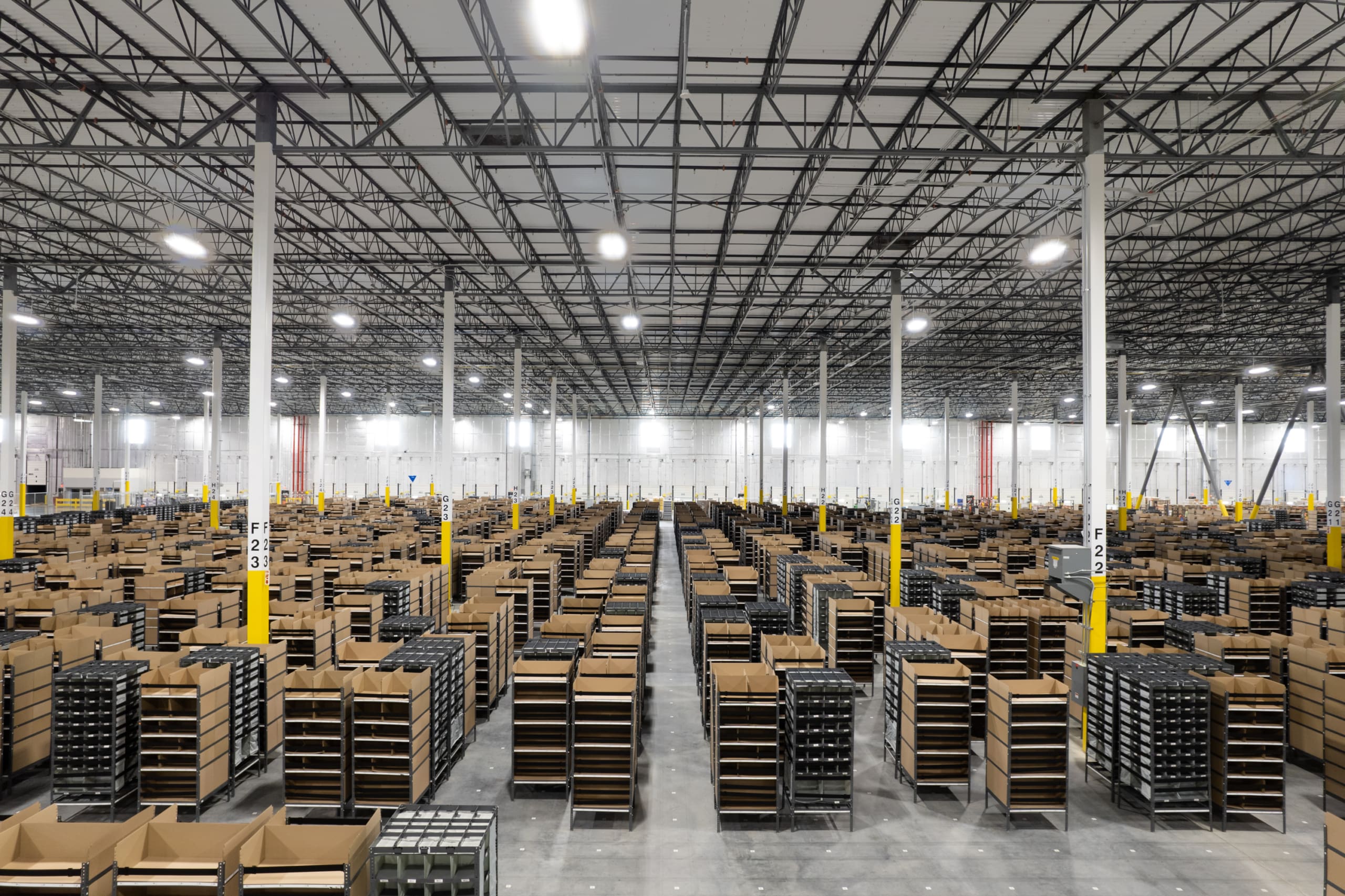 Large warehouse with numerous rows of shelves holding uniformly stacked cardboard boxes. The interior features a tall ceiling with exposed metal beams and industrial lighting.