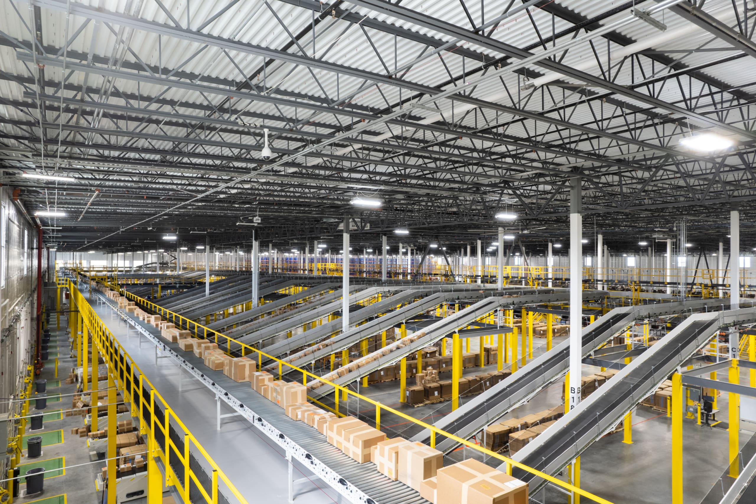 A large warehouse with numerous conveyor belts transporting boxes. The space is organized with yellow railings and high ceilings with industrial lighting.