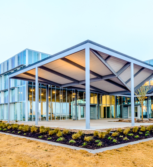 Modern glass building with geometric overhang, featuring large windows and landscaped surroundings.