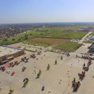 Aerial view of a large parking lot with scattered cars, adjacent to a commercial building on the left and an open field in the background. Residential houses are seen in the distance. It's a clear day.