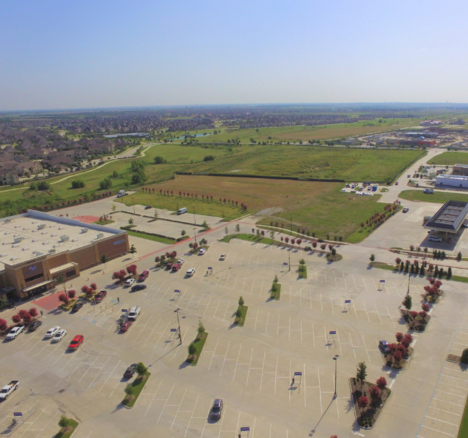 Aerial view of a large parking lot with scattered cars, adjacent to a commercial building on the left and an open field in the background. Residential houses are seen in the distance. It's a clear day.