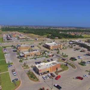 Aerial view of a commercial shopping area featuring several buildings, parking lots with multiple parked cars, and surrounding green spaces under a clear blue sky.