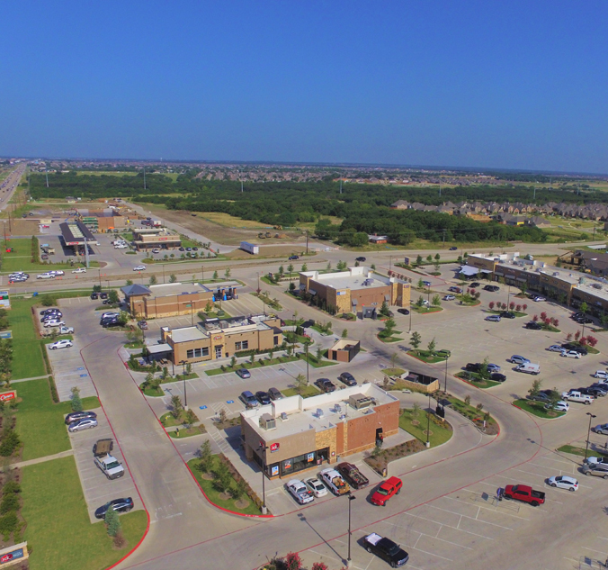 Aerial view of a commercial shopping area featuring several buildings, parking lots with multiple parked cars, and surrounding green spaces under a clear blue sky.