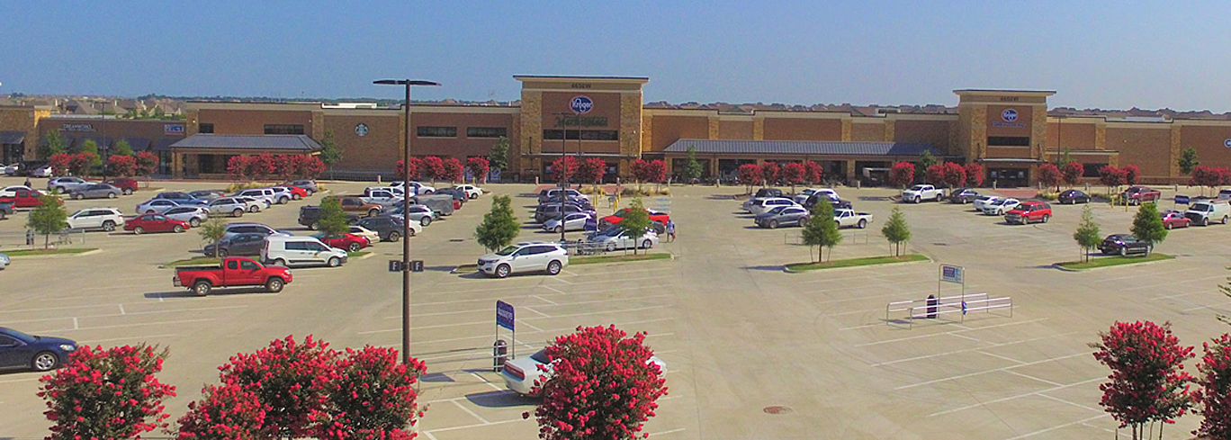 A large parking lot with numerous parked cars in front of a shopping center featuring multiple stores including a Kroger.