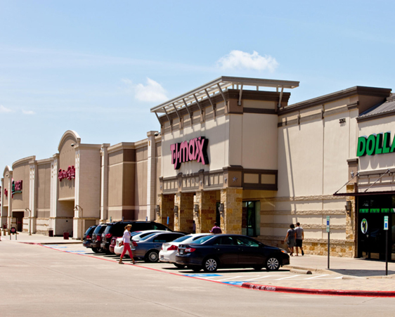 A row of retail stores on a sunny day, including a T.J. Maxx. Several cars are parked in front, and a few people are walking in the parking lot.