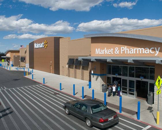 A Walmart store exterior with a sign reading "Market & Pharmacy." A car is parked near the entrance, and a few people are seen walking into the store. The sky is partly cloudy.