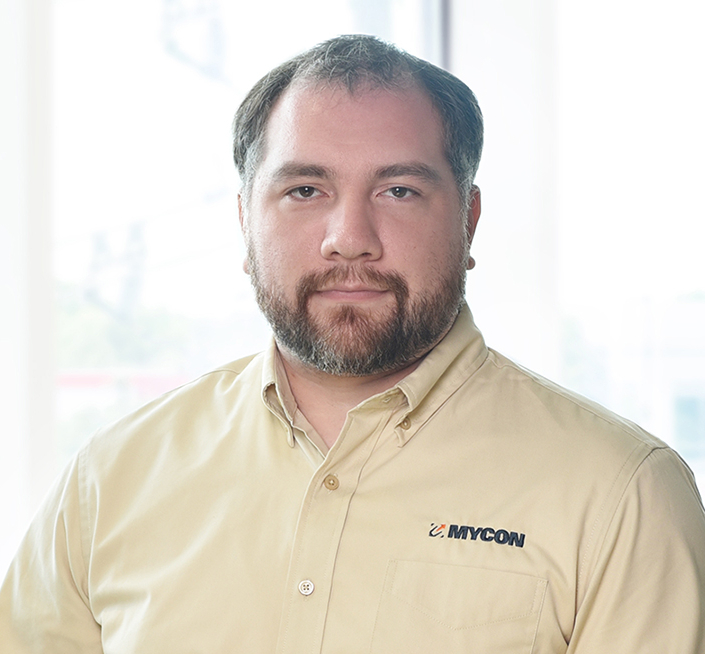 Man with short hair and a beard wearing a beige button-up shirt with the "MYCON" logo, standing indoors in front of large windows.