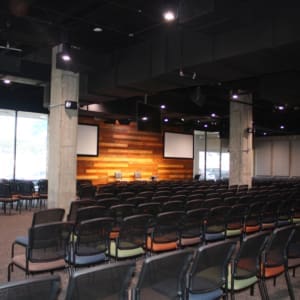 A modern conference room with rows of empty chairs facing a wooden accent wall with two screens and three chairs on a stage. Large windows line one side of the room.