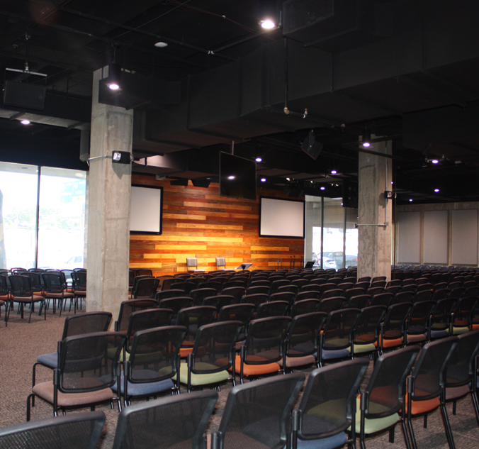 A modern conference room with rows of empty chairs facing a wooden accent wall with two screens and three chairs on a stage. Large windows line one side of the room.