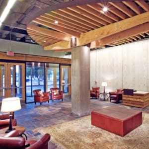 A modern lobby area featuring leather chairs, a wooden ottoman, and a rug. The space has a mix of concrete and wood elements, with large windows providing a view of trees outside.