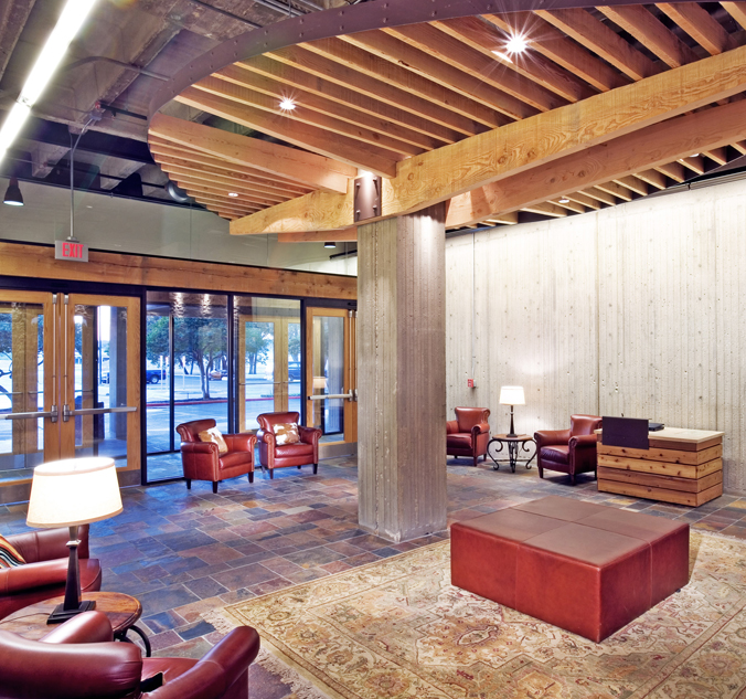 A modern lobby area featuring leather chairs, a wooden ottoman, and a rug. The space has a mix of concrete and wood elements, with large windows providing a view of trees outside.