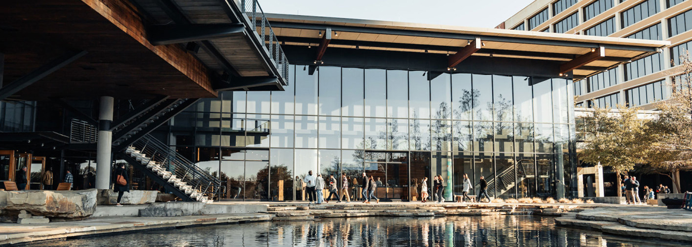 A modern glass building with a pond in front. The building has a metal overhang, and people are walking along the pathway by the water. Office structure in the background.